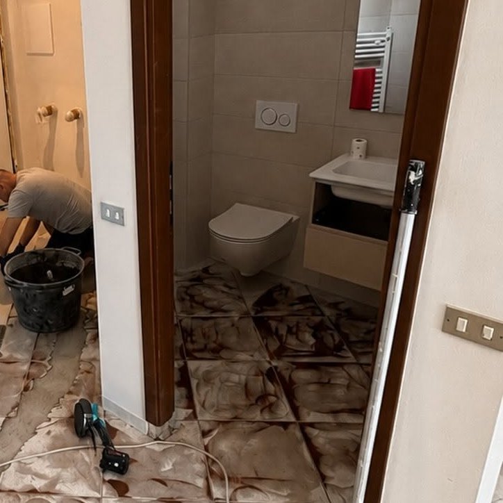 Bagno con wc sospeso e lavabo su mobile chiaro, piastrelle con residui di stucco e operaio accanto a un secchio nero